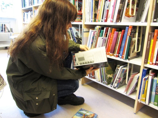 Ung flicka sitter på huk vid en bokhylla och läser en bok på ett bibliotek.