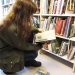 Ung flicka sitter på huk vid en bokhylla och läser en bok på ett bibliotek.